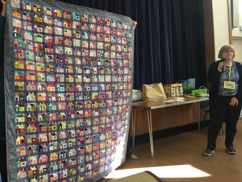 Woman with microphone speaking, whilst stood next to people holding up quilt made of squares with multicoloured houses