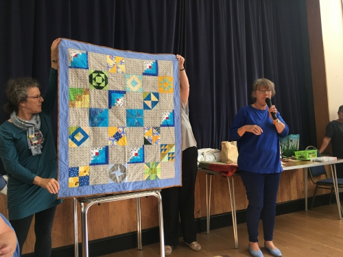 Woman with microphone speaking, whilst stood next to people holding up quilt with geometric pattern in blues, yellows and grey