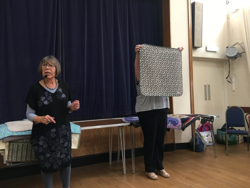 2 women standing in front of crowd. 1 holding up a grey and white quilt.