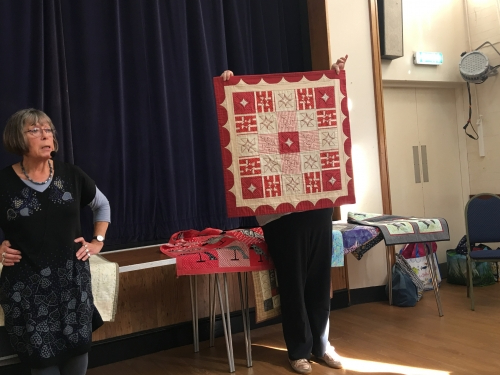 2 women standing in front of crowd. 1 holding up a quilt with a red and white block pattern