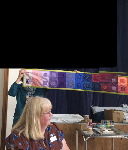 People holding end of very long and narrow quilt with multicoloured square designs