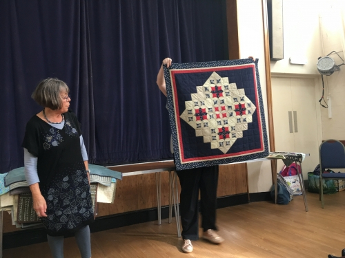 2 women standing in front of crowd. 1 holding up a red white and blue quilt.