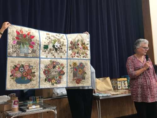 Woman with microphone speaking, whilst stood next to people holding up quilt made of 6 squares with floral design