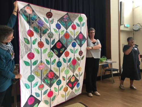Woman with microphone speaking, whilst stood next to people holding up quilt with geometric flowers and leaves