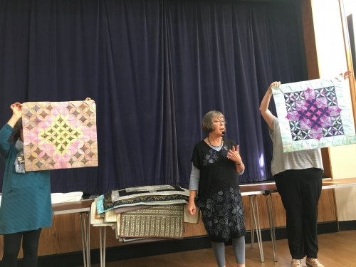 Woman speaking to crowd with microphone, with two people holding up quilts