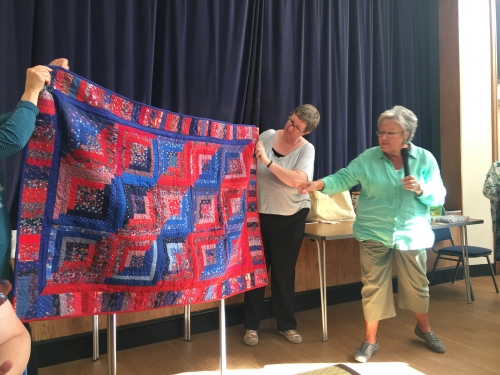 Woman with microphone speaking, whilst stood next to people holding up quilt with red and blue design