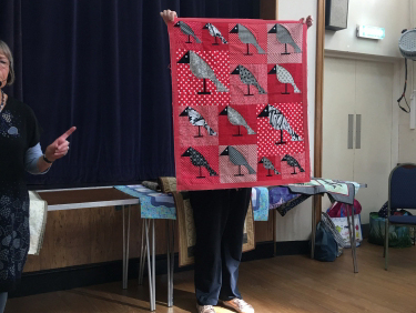 2 women standing in front of crowd. 1 holding up a quilt with a design made of multiple grey birds.