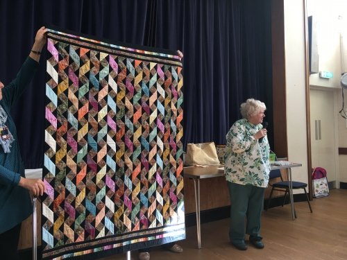 Woman with microphone speaking, whilst stood next to people holding up patterned quilt with mulit-coloured diamonds