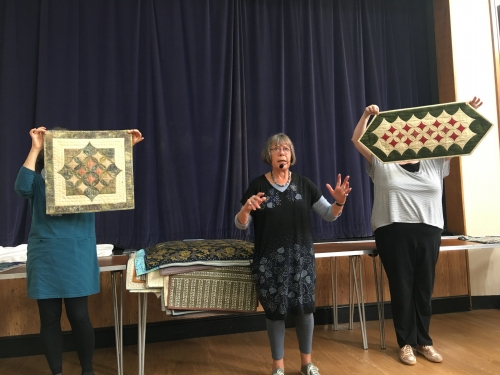 Woman speaking to crowd with microphone, with two people holding up quilts