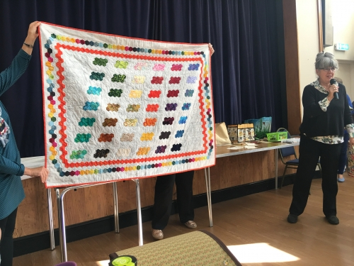 Woman with microphone speaking, whilst stood next to people holding up patterned quilt with rainbow coloured hexagons