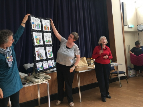 Woman with microphone stood next to women holding up quilt depicting a cartoon strip