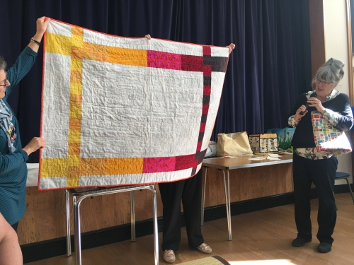 Woman with microphone speaking, whilst stood next to people holding up a white quilt with pink and yellow border