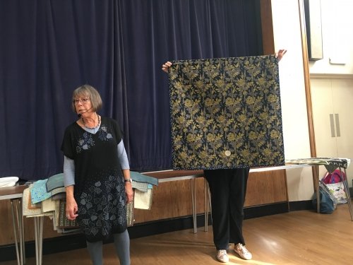 2 women standing in front of crowd. 1 holding up a black and gold quilt.