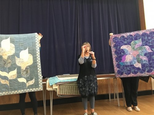 Woman speaking to crowd with microphone, with two people holding up quilts