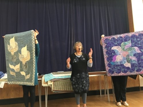 Woman speaking to crowd with microphone, with two people holding up quilts