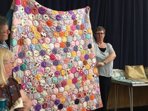 Women holding up large quilt made of multi-coloured hexagonal pieces