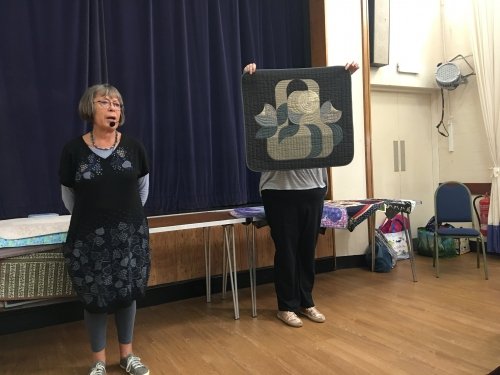 2 women standing in front of crowd. 1 holding up a black and grey quilt.