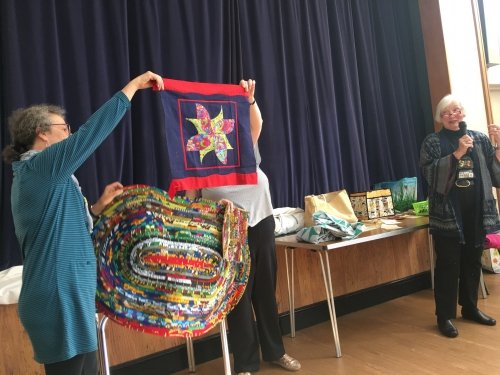 Woman speaking in microphone stood next to people holding eliptical quilt and small blue quilt with pink and green windmill pattern