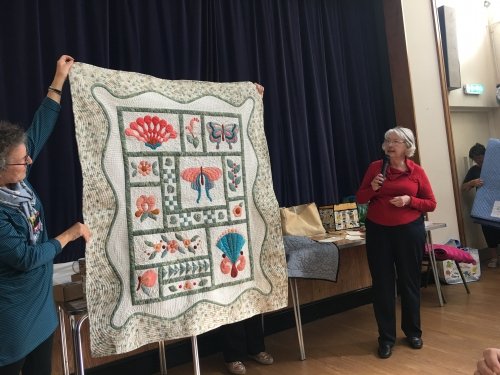 Woman with microphone next to people holding up green and white quilt with pink details