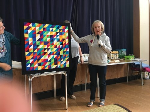 Woman with microphone speaking, whilst stood next to people holding up multi-coloured triangle patterned quilt