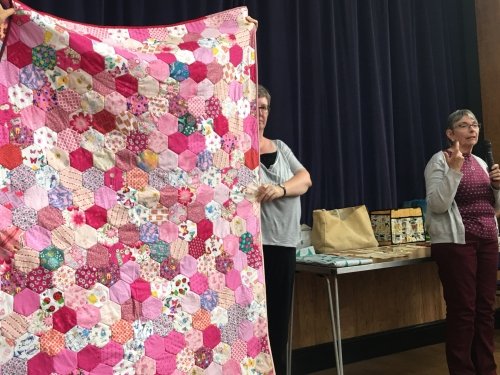 Women holding up large quilt made of multi-coloured hexagonal pieces showing pink back