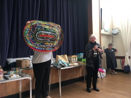 Woman with microphone speaking, whilst stood next to people holding up eliptical quilt