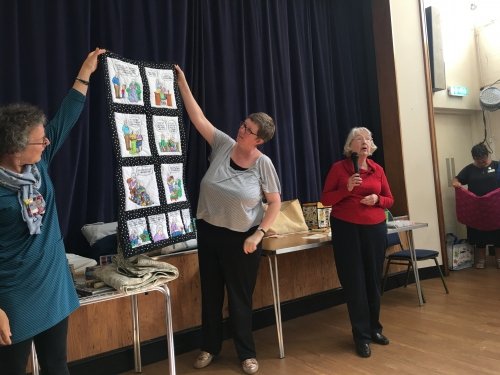 Woman with microphone stood next to women holding up quilt depicting a cartoon strip