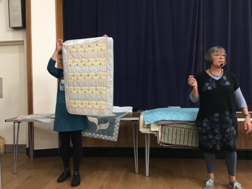 2 women standing in front of crowd. 1 holding up a pastel coloured quilt.