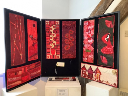 A display board showing 8 small rectangular quilts with different designs of multiple shades of red fabric