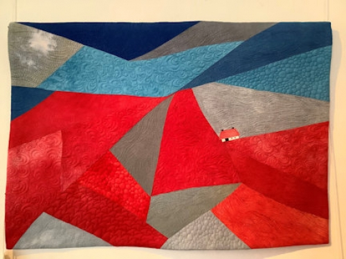 Abstract quilt made of asymmetric shaped blocks of reds, greys and blue with a small house on the edge of one shape