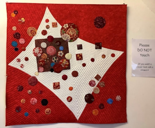 A square red quilt with an abstract cream rectangle in the centre, with an overlaid pattern of multicoloured buttons.