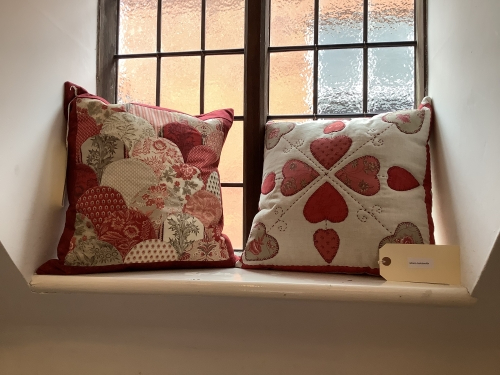 2 cushions on a window sill with red and white themed quilted cushion covers. One with a semi circle scalloped pattern, one with a hearts pattern.