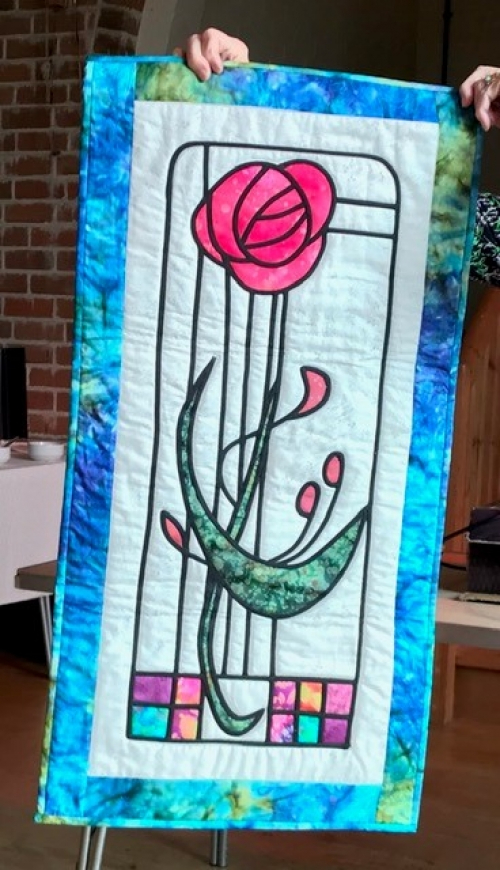 Rectangular quilt with blue border and white background depicting a contemporary style pink flower with green leaves