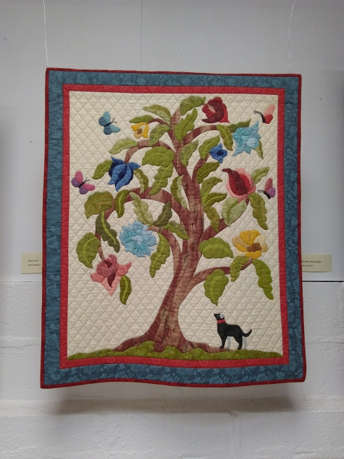 The Glory of the Garden by Jean Stetson: Large quilt depicting a tree in the centre with a dog by it's trunk looking upwards with red and blue border 