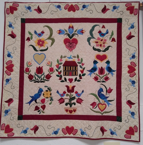 Fracture Mania by Barbara Janssen: Square quilt with house at centre and repeating pattern of blue birds and red hearts and flowers around the edge.