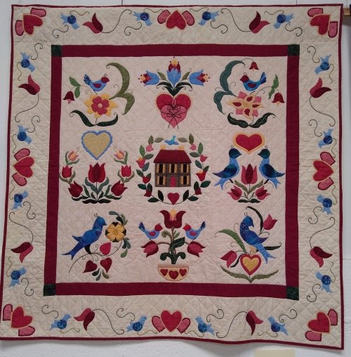 Fracture Mania by Barbara Janssen: Square quilt with house at centre and repeating pattern of blue birds and red hearts and flowers around the edge.