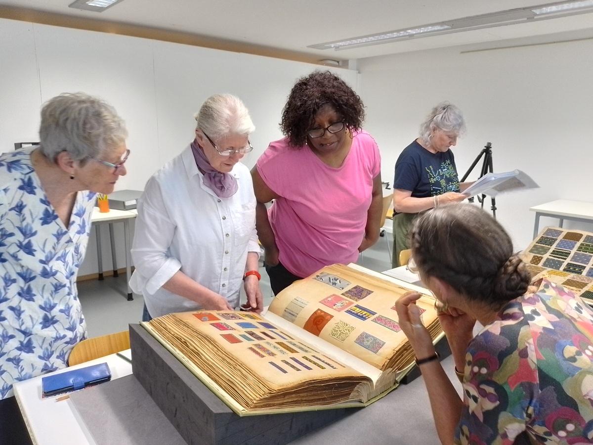 Several women studying large, old book filled with fabric samples