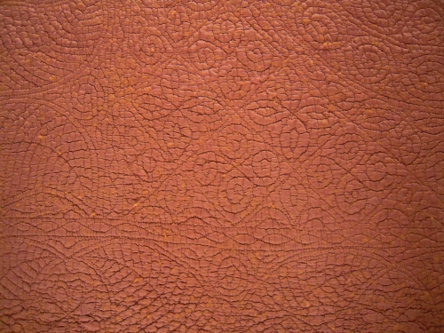 Close up of rust coloured welsh quilt showing stitching detail of squares on point with alternate rows of a spiral and leaf filling