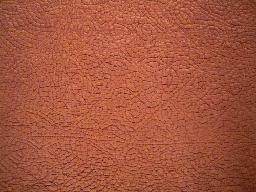 Close up of rust coloured welsh quilt showing stitching detail of squares on point with alternate rows of a spiral and leaf filling