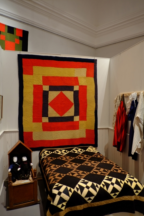 Orange, yellow and black geometric quilt hanging on wall behind bed with black and cream block quilt