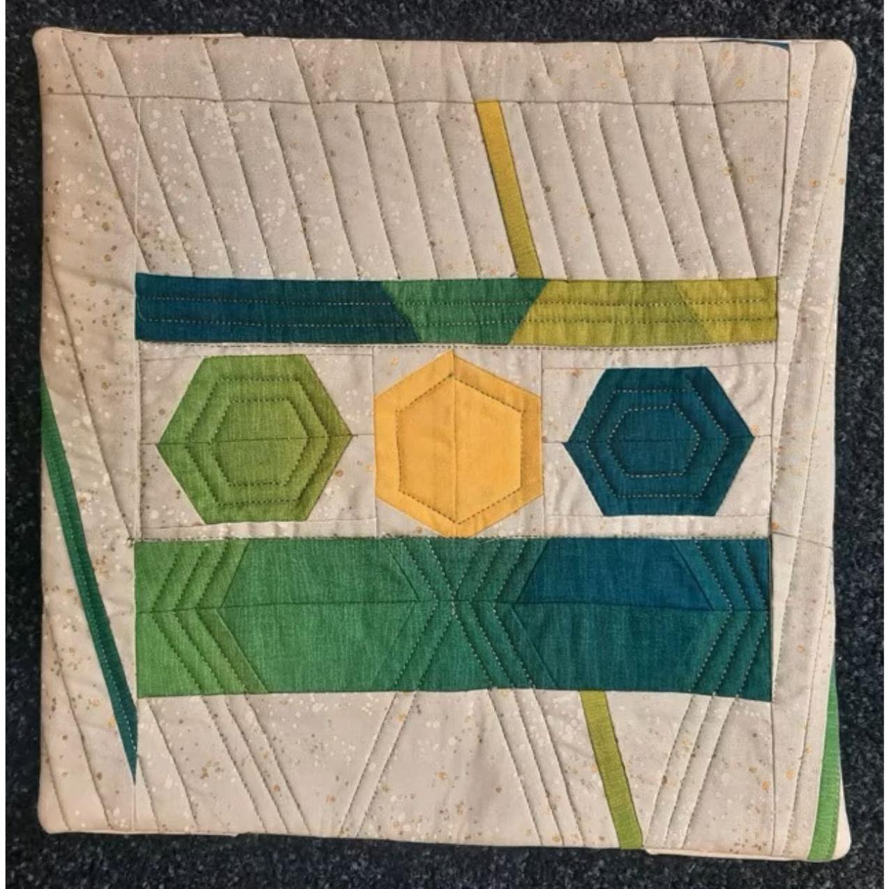 A square quilt with a beige background. The quilt features a geometric design with hexagons in shades of green and yellow. The background is quilted with diagonal lines.