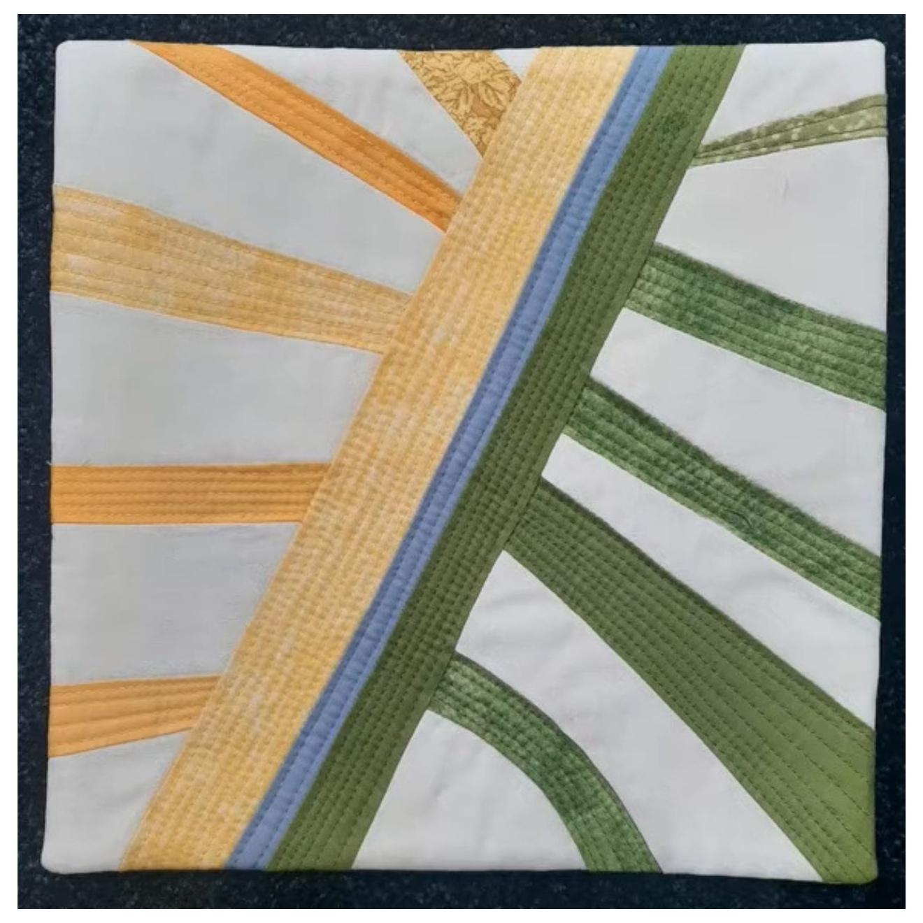 A square quilt with a white background. The quilt features a colorful, abstract design with intersecting lines and shapes in shades of green, yellow, and blue. The lines create a sense of movement and dynamism. The background is quilted with a linear pattern.