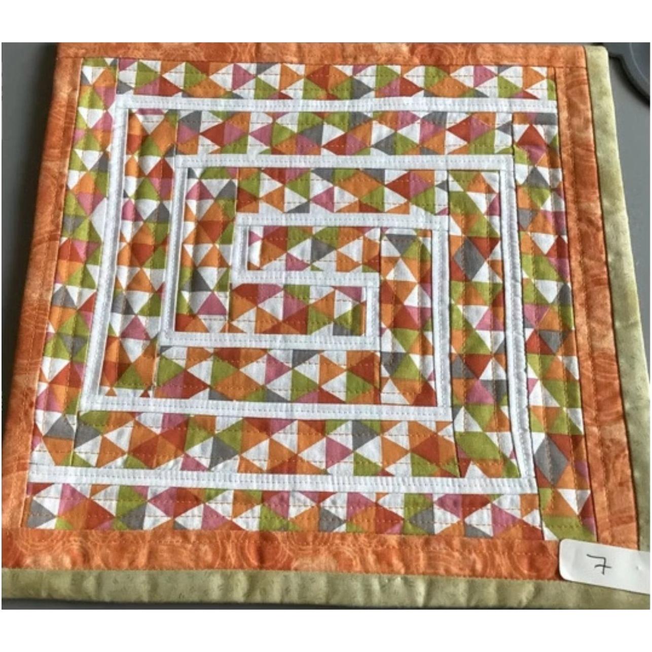 A square quilt with an orange border. The quilt features a colorful, geometric design with triangles in shades of pink, orange, green, and white. The triangles are arranged in a maze-like pattern, creating a dynamic and visually interesting composition. The background is quilted with a linear pattern.