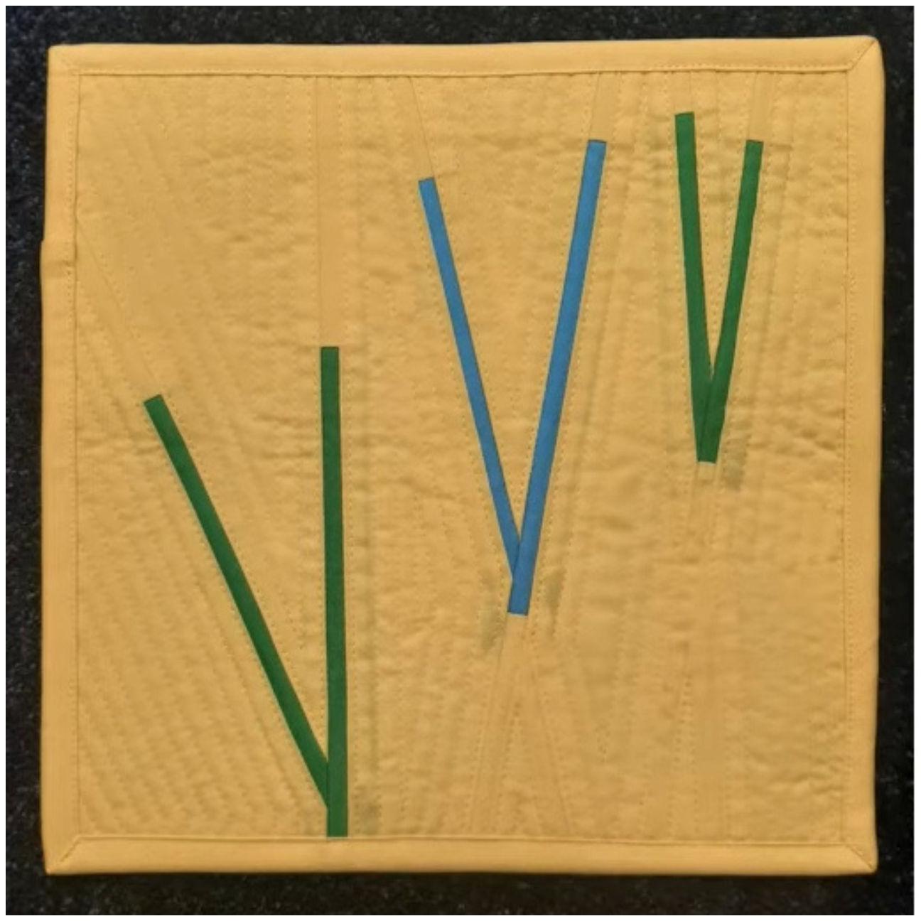 A square quilt with a yellow background. The quilt features a simple design with several green and blue vertical lines radiating outwards from the center. The background is quilted with a linear pattern.