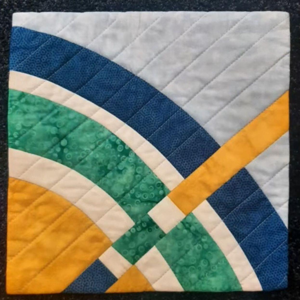 A square quilt with a blue background. The quilt features a colorful, abstract design with curved shapes in blue, green, yellow, and white. The background is quilted with a linear pattern.