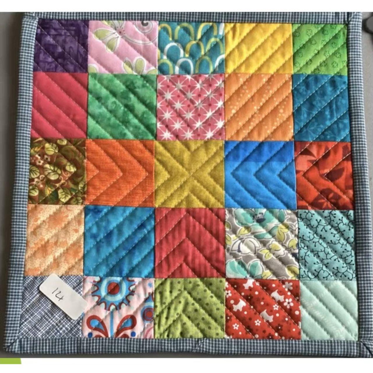 A square quilt with a patchwork design. The quilt is made up of small squares of fabric in various colors and patterns, including floral, geometric, and solid colors. The squares are arranged in a grid pattern and quilted with a diagonal line pattern. The quilt has a blue checkered border.