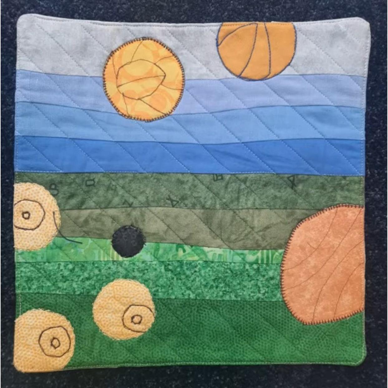 A square quilt with a landscape-like design. The top section is a blue sky with three orange circles representing the sun. The middle section is a green field with three orange circles representing pumpkins. The background is quilted with a linear pattern.