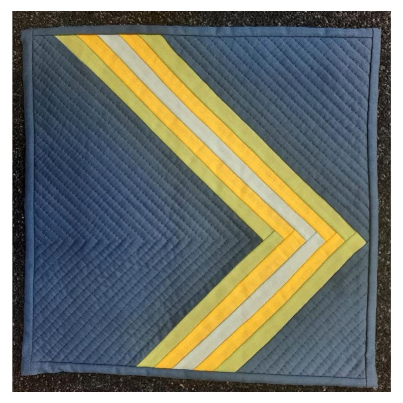 A square quilt with a dark blue background and a yellow and gray chevron pattern. The background is quilted with a subtle, linear pattern.