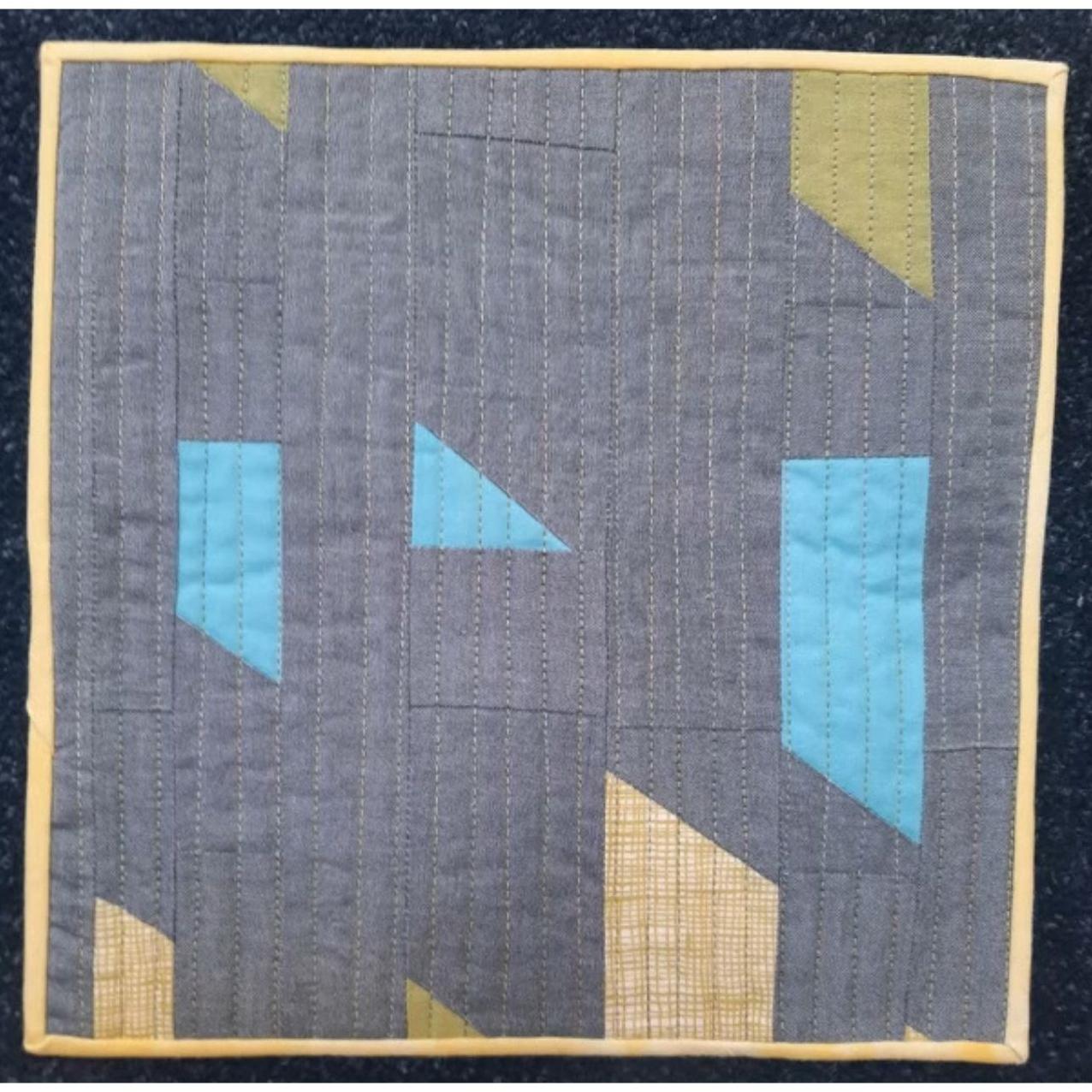 A square quilt with a gray background and a yellow border. The quilt features a geometric design with rectangles and triangles in shades of blue and green. The background is quilted with vertical lines.
