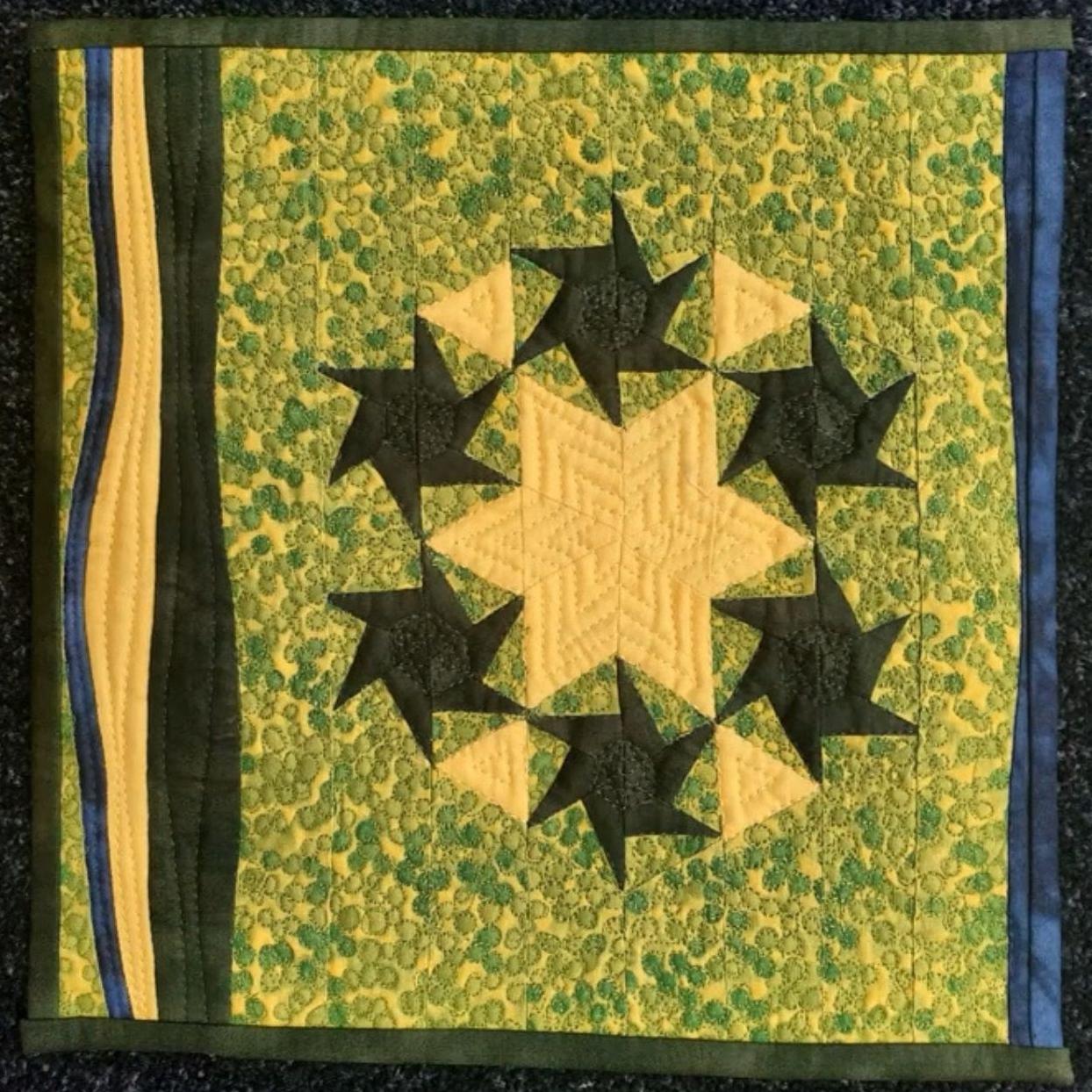  A square quilt with a green background. The quilt features a circular design with multiple black stars overlapping each other. The background is filled with a pattern of small, green shapes. The quilt has a black border with blue strips on the sides.